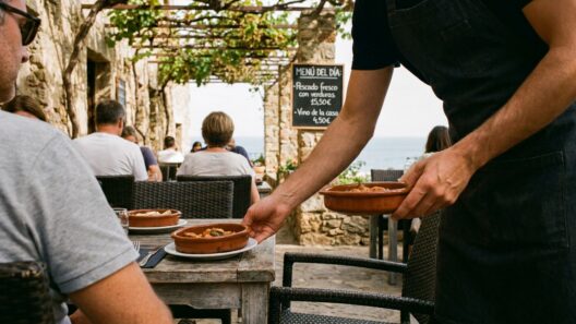 Camarero sirviendo platos en la terraza de un restaurante informal con mesas de madera en un entorno costero soleado